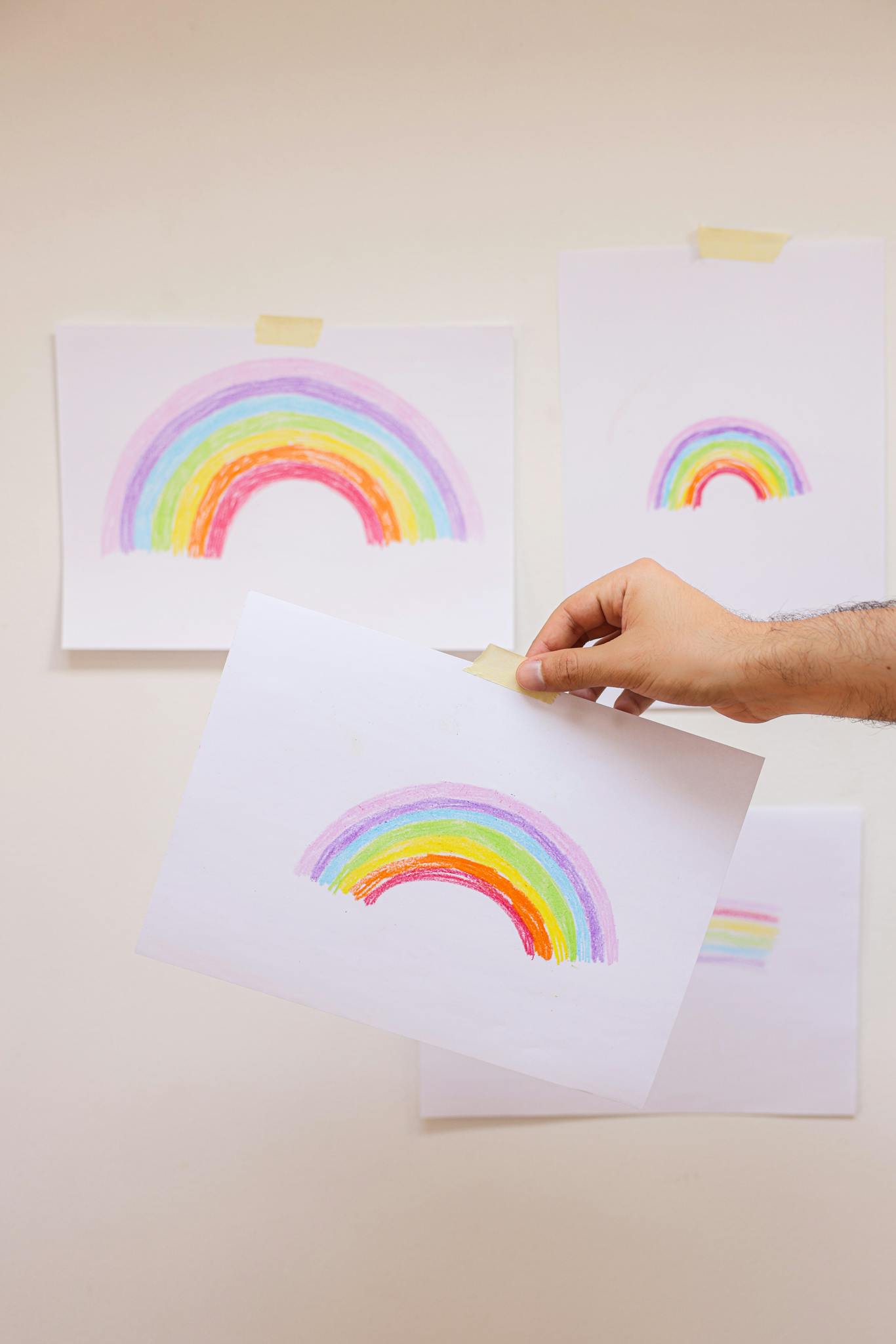 Hand holding a hand-drawn rainbow on white paper against a wall with multiple rainbow drawings taped on.